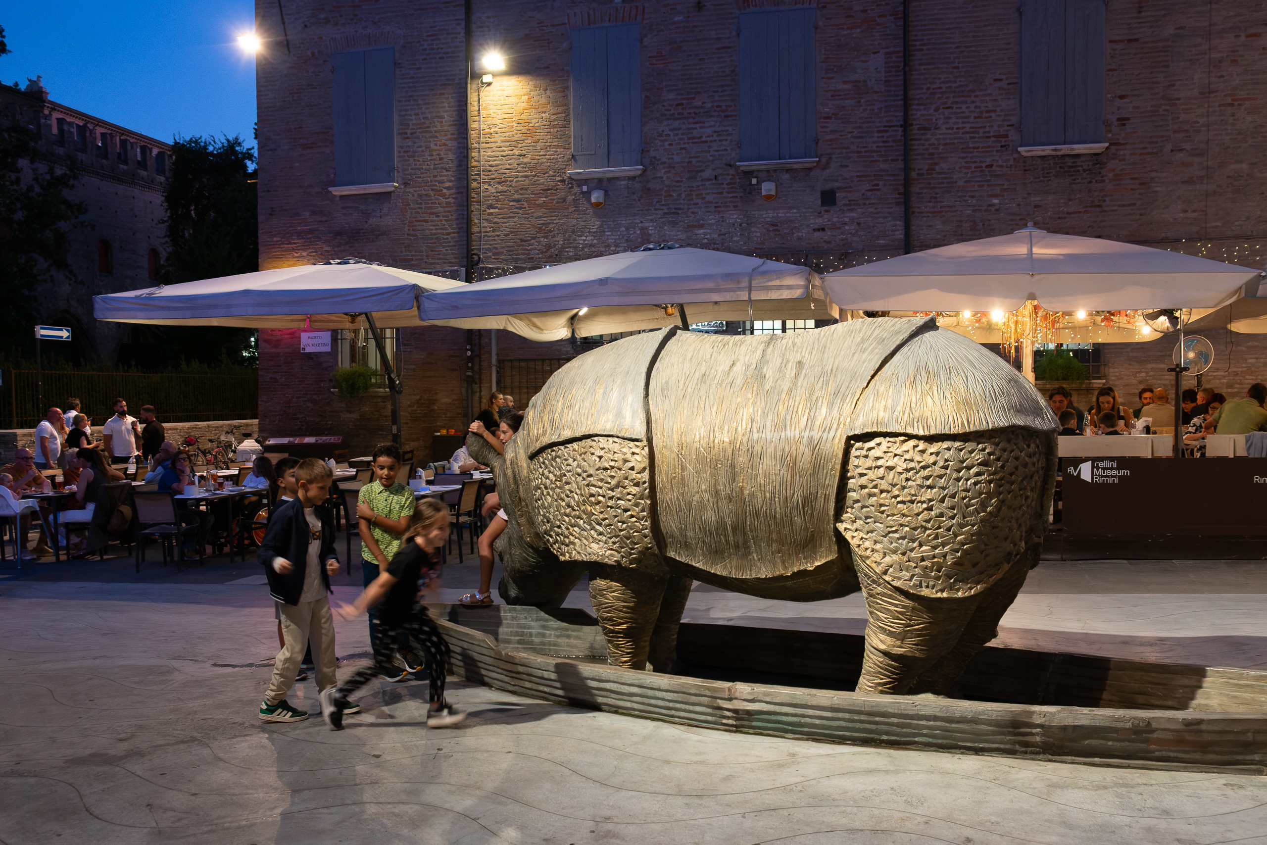 Bronze-Nashorn vor dem Fellini Museum – Kinder spielen am Brunnen